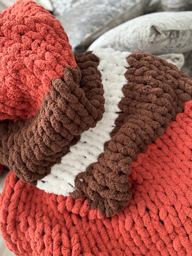 K&ouml;nnte beinhalten: Eine grob gestrickte Decke in Rost-, Braun- und Wei&szlig;t&ouml;nen. Die Decke hat dickes, strukturiertes Garn, das ein gem&uuml;tliches und einladendes Aussehen erzeugt. Das Farbschema umfasst eine rostfarbene Basis mit braunen und wei&szlig;en Streifen.