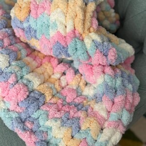 Op de afbeelding: Een zachte, handgebreide deken in pastelkleuren. De deken heeft een getextureerd, gelust ontwerp in roze, blauw, geel en wit. De gezellige deken is opgevouwen en toont het ingewikkelde steekpatroon en de kleurvariaties.