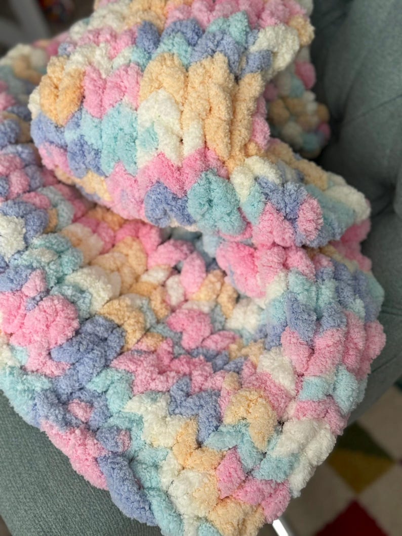 Op de afbeelding: Een zachte, handgebreide deken in pastelkleuren. De deken heeft een getextureerd, gelust ontwerp in roze, blauw, geel en wit. De gezellige deken is opgevouwen en toont het ingewikkelde steekpatroon en de kleurvariaties.
