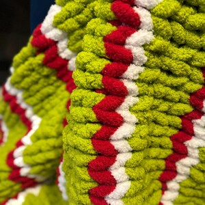May include: Close-up of a chunky knit blanket in shades of green, red, and white. The blanket features a repeating chevron pattern in red and white against a green background. The texture appears soft and plush.