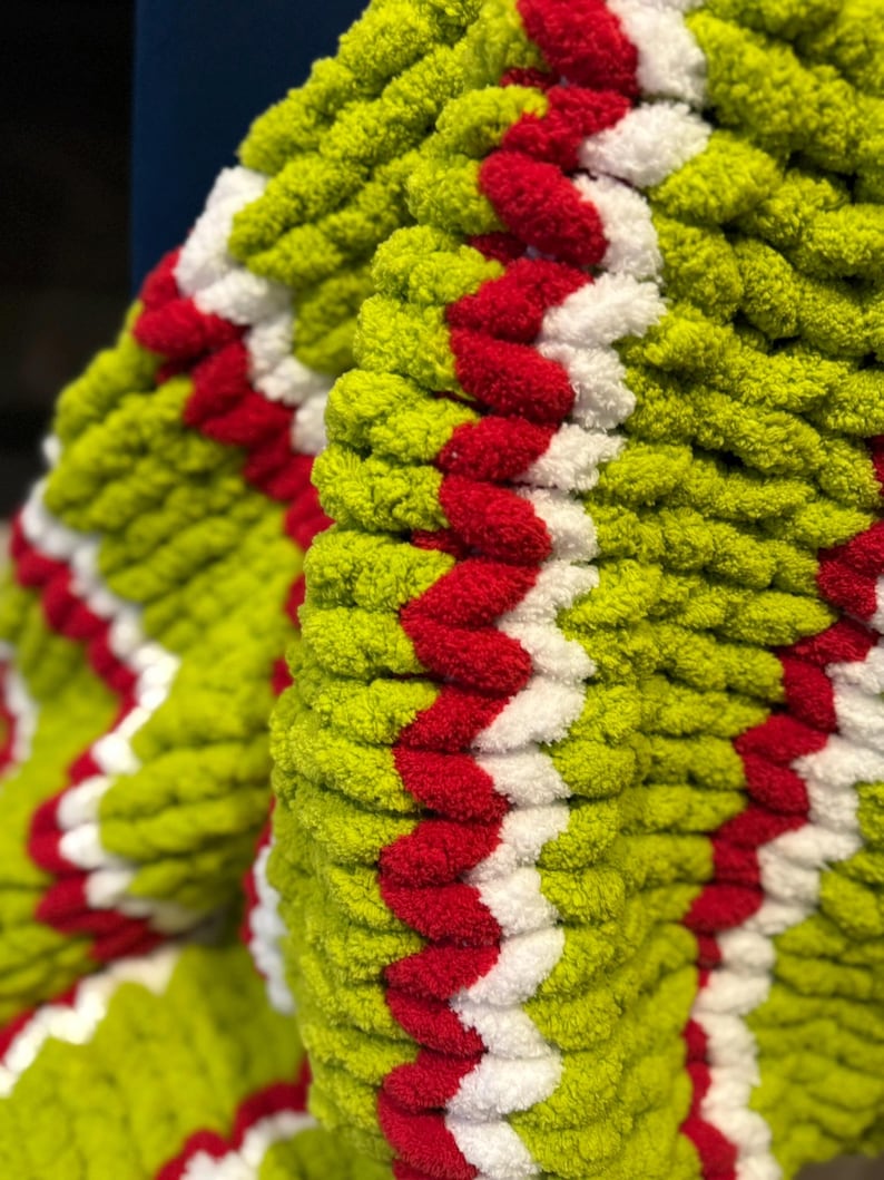 May include: Close-up of a chunky knit blanket in shades of green, red, and white. The blanket features a repeating chevron pattern in red and white against a green background. The texture appears soft and plush.