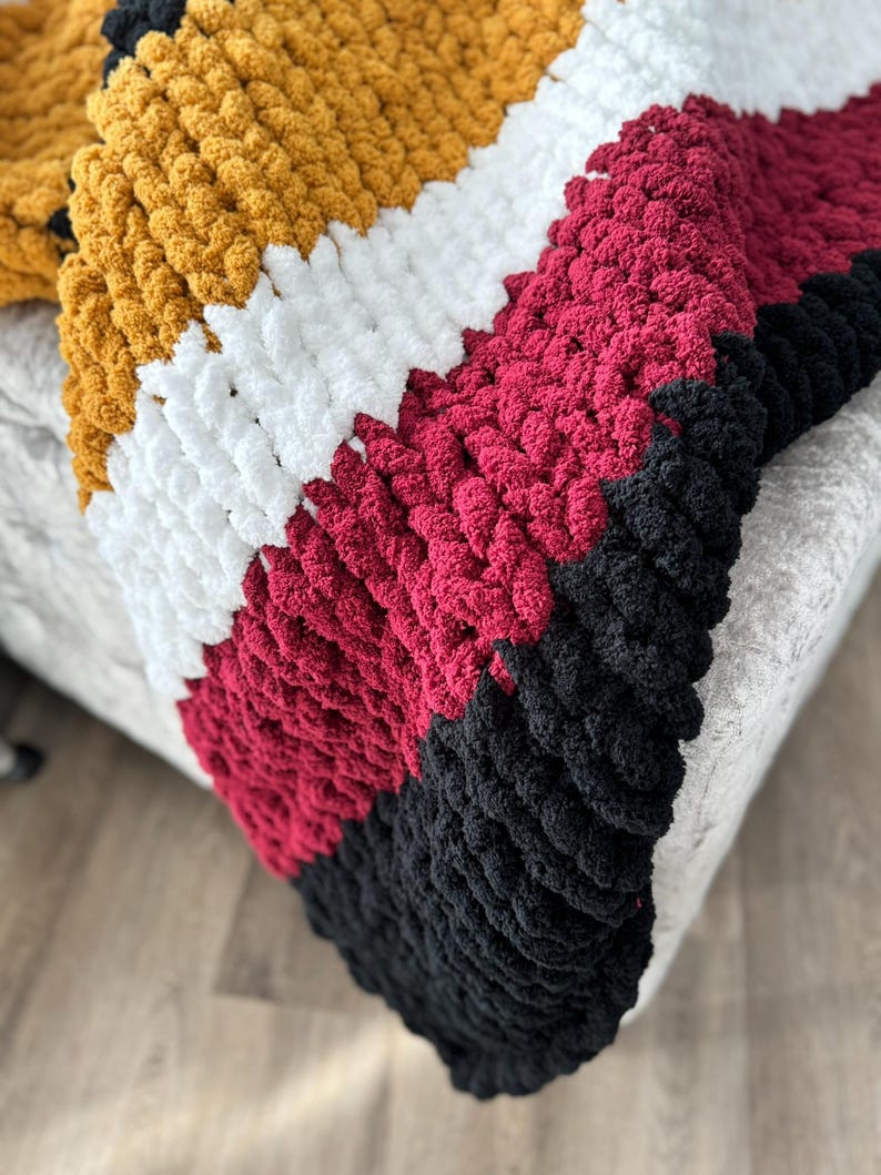 May include: A chunky knit throw blanket with stripes of mustard yellow, white, burgundy, and black yarn.