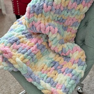 Op de afbeelding: Een pastelkleurige, dik gebreide deken in tinten roze, blauw, geel en groen. De deken is over een lichtgroene stoel gedrapeerd, wat de zachte textuur en kleurvariaties laat zien. De gezellige, handgemaakte deken is een woondecoratie-artikel.