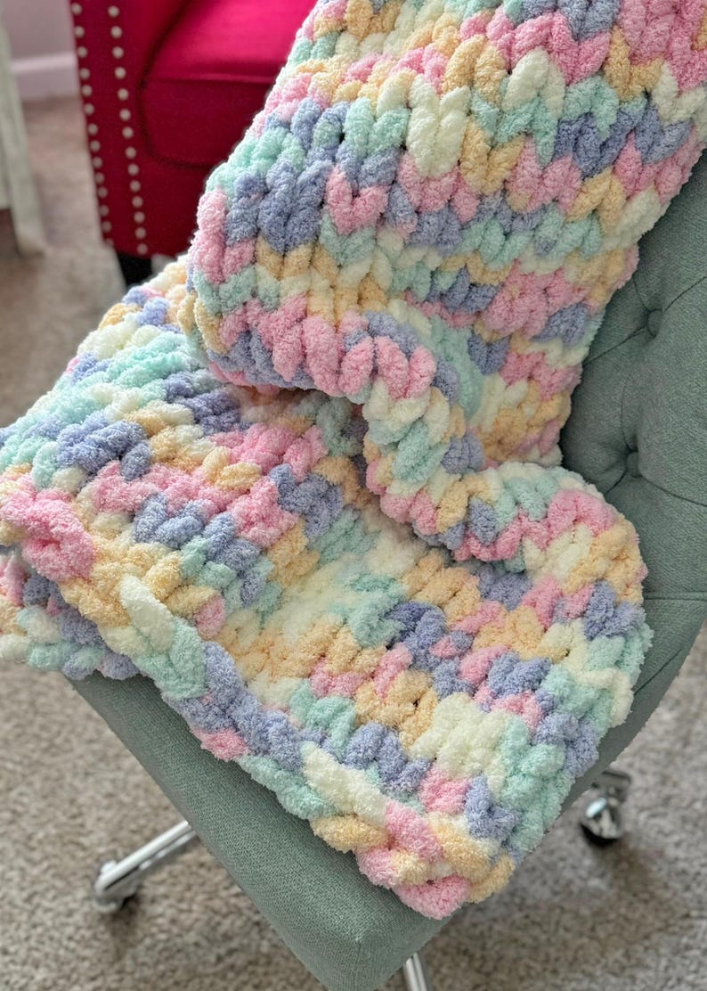 Op de afbeelding: Een pastelkleurige, dik gebreide deken in tinten roze, blauw, geel en groen. De deken is over een lichtgroene stoel gedrapeerd, wat de zachte textuur en kleurvariaties laat zien. De gezellige, handgemaakte deken is een woondecoratie-artikel.