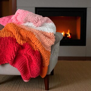 Peut inclure: Une couverture multicolore, drap&eacute;e sur un fauteuil gris. Elle pr&eacute;sente des rayures rouges, oranges, p&ecirc;che, blanches et roses. La texture douce sugg&egrave;re une sensation de chaleur et d'invitation. Un foyer avec un feu ardent est visible en arri&egrave;re-plan.