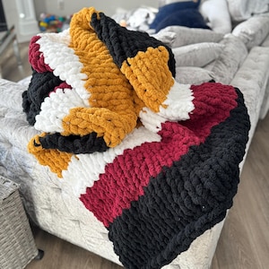 May include: A chunky knit throw blanket with stripes of black, white, burgundy, and gold yarn.