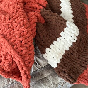 K&ouml;nnte beinhalten: Eine grob gestrickte Decke in Rost-, Braun- und Wei&szlig;t&ouml;nen. Die Decke hat ein Streifendesign mit einem rostfarbenen Abschnitt auf der linken Seite, der in braune und wei&szlig;e Streifen auf der rechten Seite &uuml;bergeht. Das dicke Garn erzeugt ein gem&uuml;tliches, strukturiertes Aussehen.