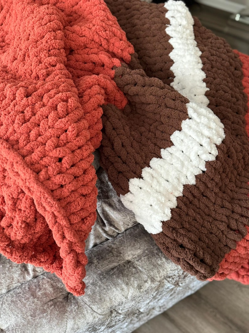K&ouml;nnte beinhalten: Eine grob gestrickte Decke in Rost-, Braun- und Wei&szlig;t&ouml;nen. Die Decke hat ein Streifendesign mit einem rostfarbenen Abschnitt auf der linken Seite, der in braune und wei&szlig;e Streifen auf der rechten Seite &uuml;bergeht. Das dicke Garn erzeugt ein gem&uuml;tliches, strukturiertes Aussehen.