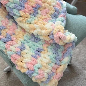 Op de afbeelding: Een handgemaakte, dik gebreide deken in pastelkleuren roze, geel, blauw en groen. De deken is over een lichtgroene stoel gedrapeerd. Het zachte, getextureerde garen cre&euml;ert een gezellige en uitnodigende uitstraling.