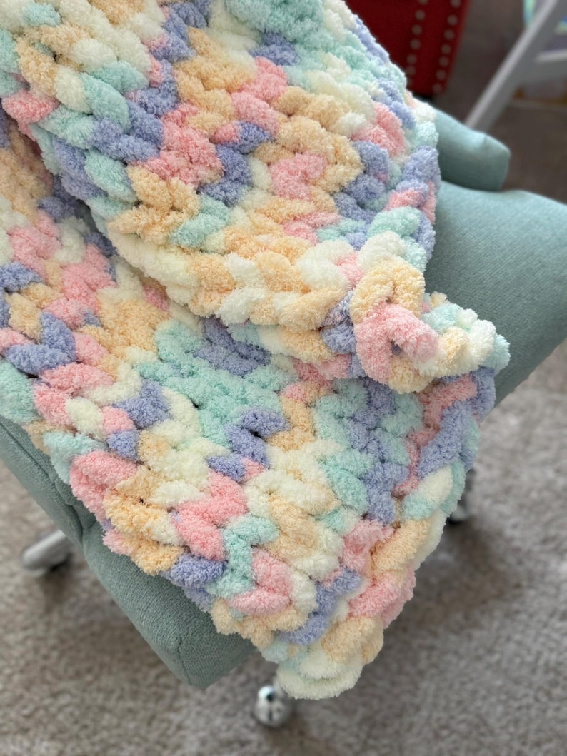 Op de afbeelding: Een handgemaakte, dik gebreide deken in pastelkleuren roze, geel, blauw en groen. De deken is over een lichtgroene stoel gedrapeerd. Het zachte, getextureerde garen cre&euml;ert een gezellige en uitnodigende uitstraling.