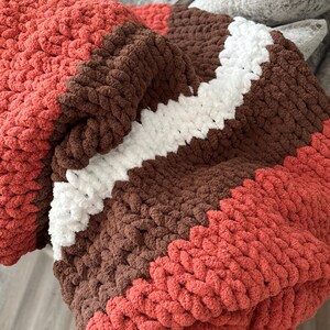 K&ouml;nnte beinhalten: Eine grob gestrickte Decke in den Farben Koralle, Braun und Wei&szlig;. Die Decke hat einen diagonalen wei&szlig;en Streifen in der Mitte, mit Koralle oben und unten. Das dicke Garn erzeugt ein gem&uuml;tliches, strukturiertes Aussehen.