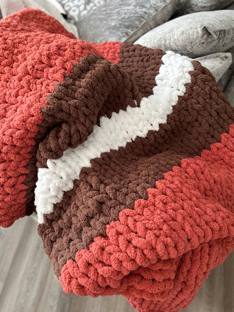K&ouml;nnte beinhalten: Eine grob gestrickte Decke in den Farben Koralle, Braun und Wei&szlig;. Die Decke hat einen diagonalen wei&szlig;en Streifen in der Mitte, mit Koralle oben und unten. Das dicke Garn erzeugt ein gem&uuml;tliches, strukturiertes Aussehen.