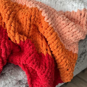 Peut inclure: Une couverture tricot&eacute;e &agrave; la main avec un design color block. La couverture pr&eacute;sente des sections rouges, oranges, p&ecirc;che, blanches et roses. La texture en tricot &eacute;pais ajoute &agrave; la chaleur et &agrave; l'attrait visuel de la couverture.