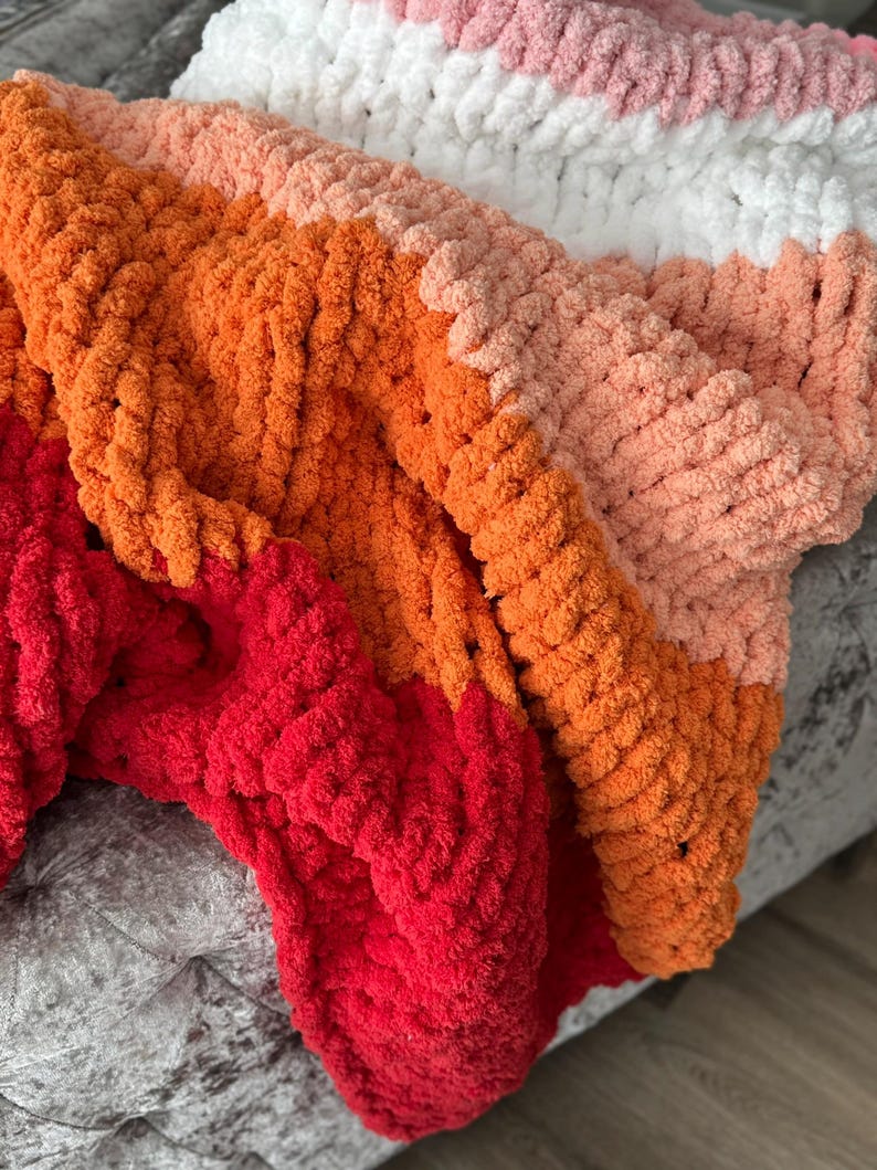 Peut inclure: Une couverture tricot&eacute;e &agrave; la main avec un design color block. La couverture pr&eacute;sente des sections rouges, oranges, p&ecirc;che, blanches et roses. La texture en tricot &eacute;pais ajoute &agrave; la chaleur et &agrave; l'attrait visuel de la couverture.