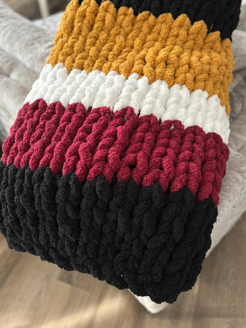 May include: A chunky knit throw blanket with stripes of mustard yellow, white, burgundy, and black yarn.