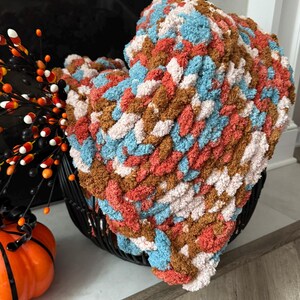 May include: A chunky hand-knit blanket featuring a variegated pattern of soft yarn in shades of light blue, cream, burnt orange, and brown. The thick, textured loops create a dense, plush appearance throughout the entire surface of the throw.