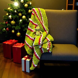May include: A green, red, and white striped knitted blanket draped over a chair, with a small plush toy. A Christmas tree with gold ornaments and red gift boxes are in the background.