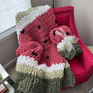 May include: A handmade watermelon-themed blanket and matching accessories. The blanket features a red center with black seed accents, white, light green, and dark green borders. The set includes two matching accessories, all made of a chunky knit material.