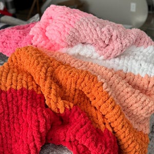 Peut inclure: Gros plan sur plusieurs couvertures en tricot &eacute;pais de diff&eacute;rentes couleurs. Les couvertures sont dans des tons de rose, d'orange et de blanc. La texture du fil est visible, cr&eacute;ant une apparence douce et confortable. Les couvertures sont pli&eacute;es et superpos&eacute;es, mettant en valeur les diff&eacute;rentes couleurs.