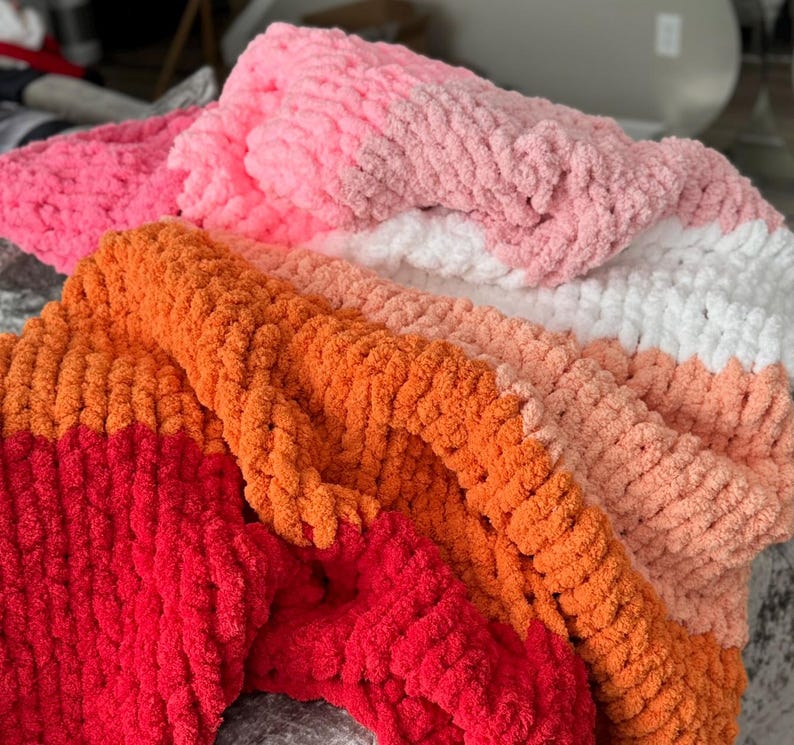 Peut inclure: Gros plan sur plusieurs couvertures en tricot &eacute;pais de diff&eacute;rentes couleurs. Les couvertures sont dans des tons de rose, d'orange et de blanc. La texture du fil est visible, cr&eacute;ant une apparence douce et confortable. Les couvertures sont pli&eacute;es et superpos&eacute;es, mettant en valeur les diff&eacute;rentes couleurs.