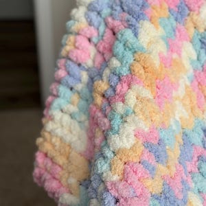 Op de afbeelding: Close-up van een zachte, dikke gebreide deken in pastelkleuren. De deken heeft een herhalend patroon van roze, blauw, geel en wit. De textuur ziet er zacht en gezellig uit.