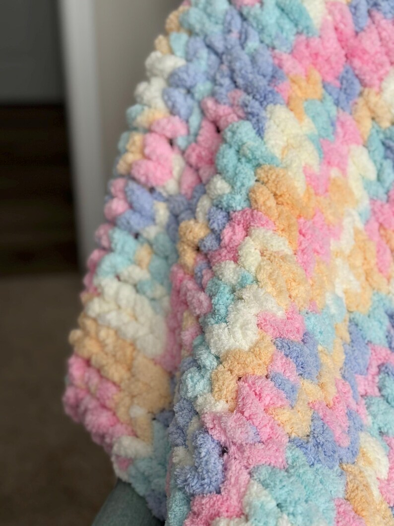 Op de afbeelding: Close-up van een zachte, dikke gebreide deken in pastelkleuren. De deken heeft een herhalend patroon van roze, blauw, geel en wit. De textuur ziet er zacht en gezellig uit.