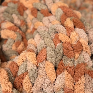 May include: A chunky knit throw blanket in shades of brown, beige, and peach. The blanket is made with a thick, textured yarn and has a soft, cozy feel.