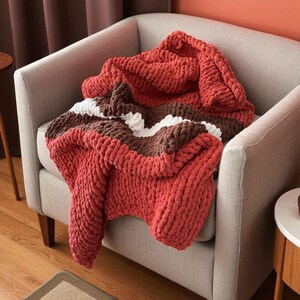 K&ouml;nnte beinhalten: Eine grob gestrickte Decke in Rot-, Braun- und Wei&szlig;t&ouml;nen liegt &uuml;ber einem grauen Sessel. Die Decke hat ein gestreiftes Design mit einem dicken, strukturierten Aussehen, das eine gem&uuml;tliche und einladende &Auml;sthetik schafft. Die warmen Farben und die weiche Textur suggerieren Komfort.