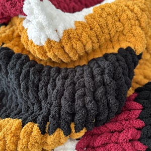 May include: A close-up of a chunky knit blanket with stripes of black, white, burgundy, and mustard yellow yarn.