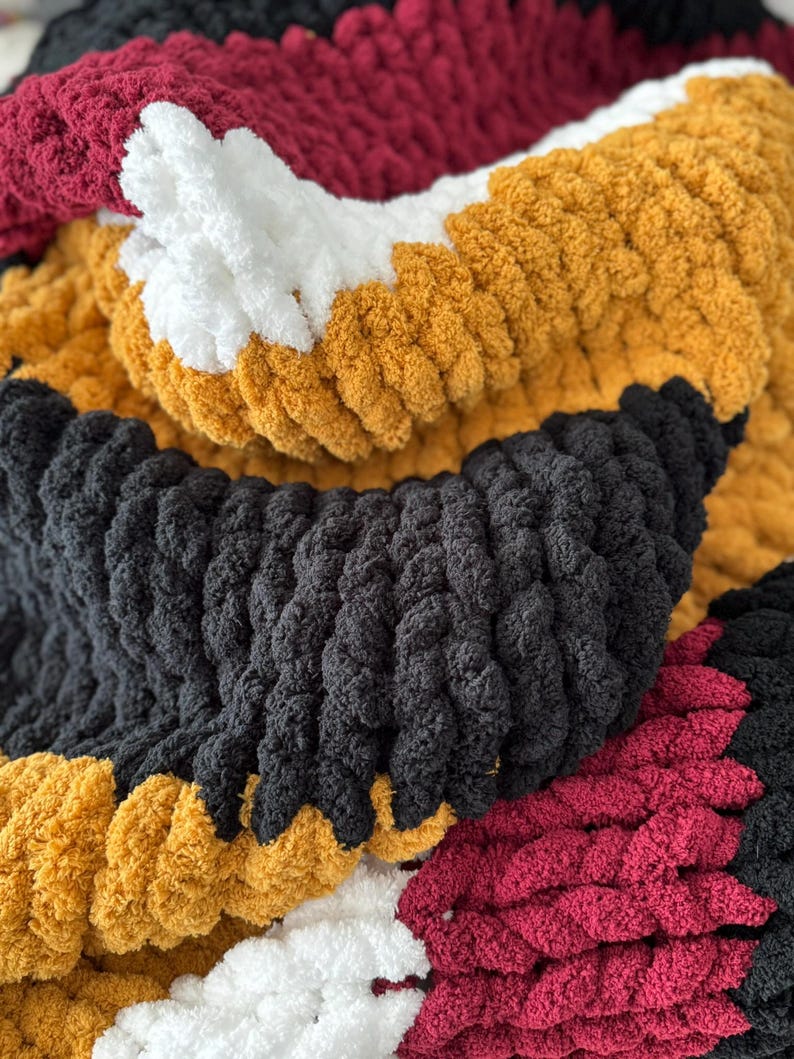 May include: A close-up of a chunky knit blanket with stripes of black, white, burgundy, and mustard yellow yarn.