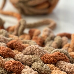 K&ouml;nnte beinhalten: Nahaufnahme einer dicken Strickdecke in Braun-, Beige- und Gr&uuml;nt&ouml;nen. Die Decke ist aus dickem Garn gefertigt und hat eine weiche, flauschige Textur.
