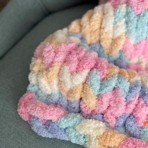 Op de afbeelding: Close-up van een dikke gebreide deken in pastelkleuren. De deken heeft een gevlochten ontwerp met tinten roze, blauw, geel en wit. De zachte, donzige textuur is zichtbaar, wat duidt op een gezellig en comfortabel item.