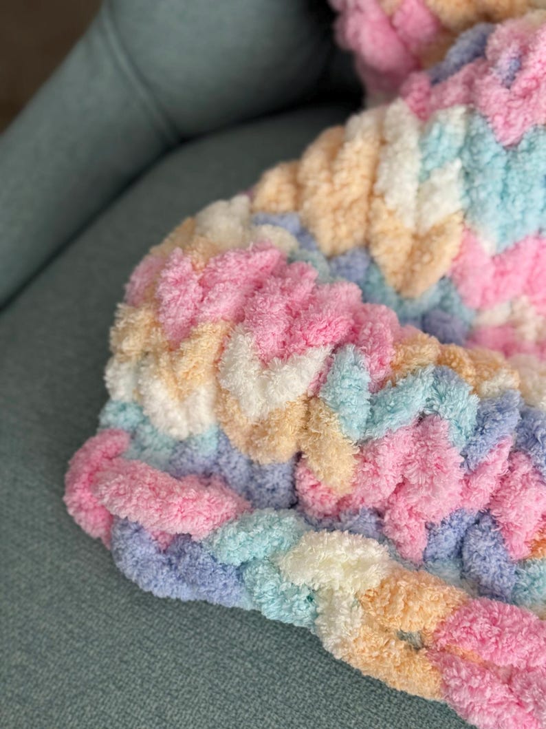 Op de afbeelding: Close-up van een dikke gebreide deken in pastelkleuren. De deken heeft een gevlochten ontwerp met tinten roze, blauw, geel en wit. De zachte, donzige textuur is zichtbaar, wat duidt op een gezellig en comfortabel item.