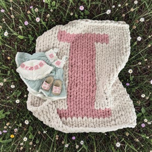May include: A beige and pink chunky knit baby blanket with a large pink letter "I". A matching outfit including a hat, top, shorts, and shoes is next to the blanket. The outfit is light blue with white and pink accents.