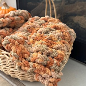 K&ouml;nnte beinhalten: Ein geflochtener Korb gef&uuml;llt mit einer dicken Strickdecke in Braun-, Orange- und Beiget&ouml;nen. Die Decke ist aus weichem, flauschigem Garn gefertigt und hat ein strukturiertes, Zopfmuster.