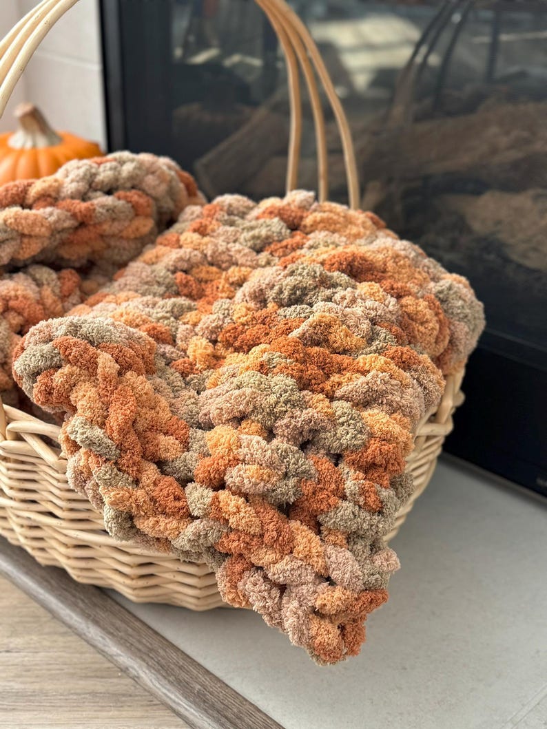 K&ouml;nnte beinhalten: Ein geflochtener Korb gef&uuml;llt mit einer dicken Strickdecke in Braun-, Orange- und Beiget&ouml;nen. Die Decke ist aus weichem, flauschigem Garn gefertigt und hat ein strukturiertes, Zopfmuster.