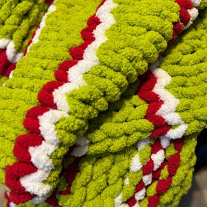 May include: A close-up of a chunky knit blanket in shades of green, red, and white. The blanket features a zig-zag pattern with a vibrant green base, accented by bold red and white stripes. The thick, textured yarn creates a cozy, inviting look.