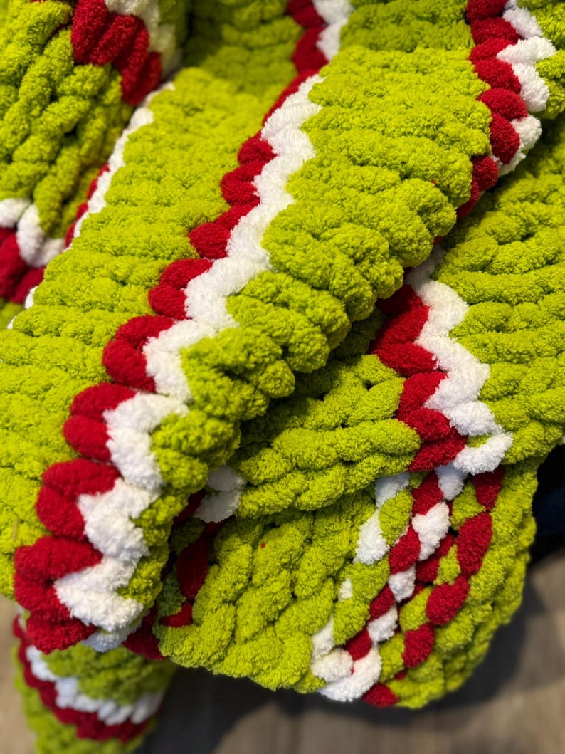 May include: A close-up of a chunky knit blanket in shades of green, red, and white. The blanket features a zig-zag pattern with a vibrant green base, accented by bold red and white stripes. The thick, textured yarn creates a cozy, inviting look.