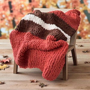 K&ouml;nnte beinhalten: Eine grob gestrickte Decke in Rot-, Braun- und Wei&szlig;t&ouml;nen ist &uuml;ber einen Holzstuhl drapiert. Die Decke hat ein gestreiftes Design und ein dickes, strukturiertes Gewebe, perfekt f&uuml;r gem&uuml;tlichen Komfort. Die Decke besteht aus einem weichen, pl&uuml;schigen Material.
