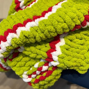 May include: A close-up of a hand-knitted blanket. The blanket is a vibrant lime green with a zig-zag pattern of red and white. The chunky knit creates a textured, cozy appearance, perfect for home decor.