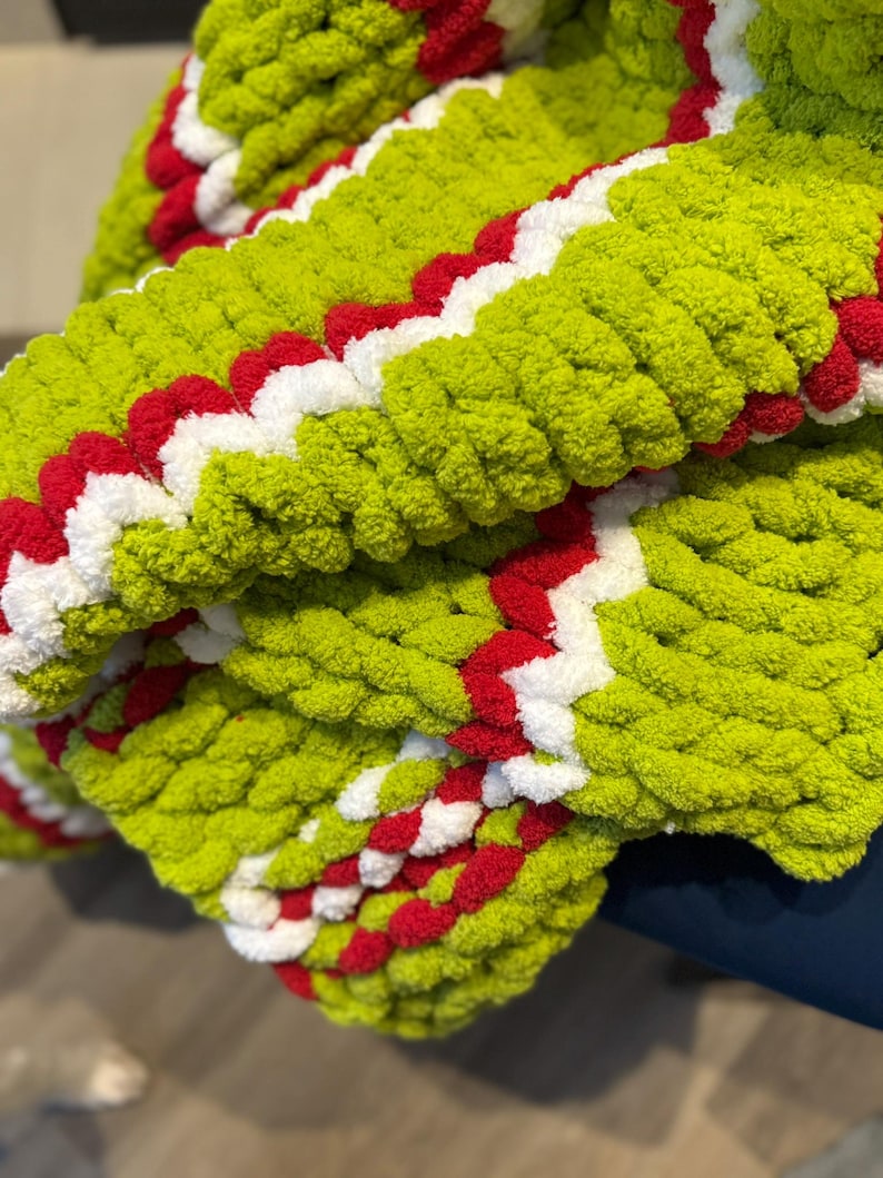 May include: A close-up of a hand-knitted blanket. The blanket is a vibrant lime green with a zig-zag pattern of red and white. The chunky knit creates a textured, cozy appearance, perfect for home decor.