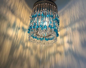 May include: A chandelier with a round, tiered shade made of clear and turquoise glass teardrop crystals. The chandelier is hanging from a ceiling.