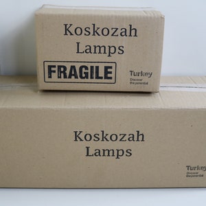 May include: Two brown cardboard boxes stacked on top of each other. The top box has the text "Koskozah Lamps" and "FRAGILE" printed on it. The bottom box has the text "Koskozah Lamps" printed on it. Both boxes have a sticker that says "Turkey Discover the potential."
