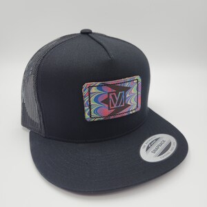 May include: Black trucker hat with a black mesh back. The hat has a square patch with a colorful swirl pattern and a white letter "M" inside.