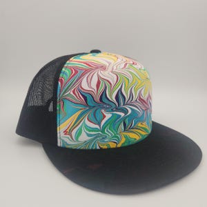 May include: A black and white trucker hat with a colourful marbled design on the front. The hat has a black mesh back and a black brim.