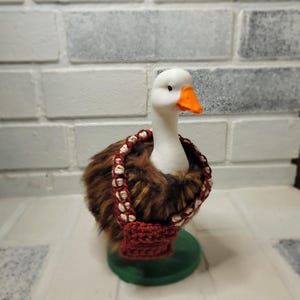May include: A decorative goose figurine with a white head, orange beak, and brown and tan body. It wears a red crocheted purse with a white and red strap. The goose is mounted on a green base.