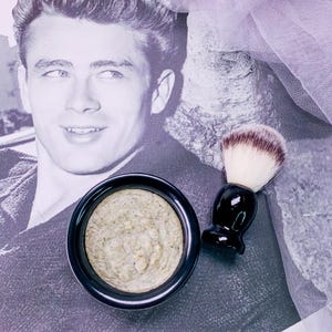May include: A black bowl filled with a light beige substance, next to a black and white shaving brush. The background features a black and white portrait of a man.