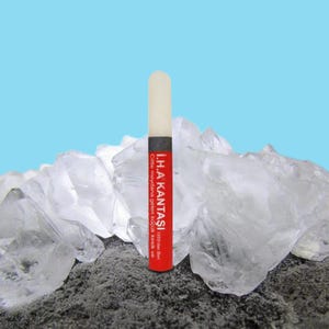 May include: A red and white rectangular object with text, a black band, and a white top. The object is standing on a bed of gray material, with large, clear, crystalline structures in the background against a blue sky.