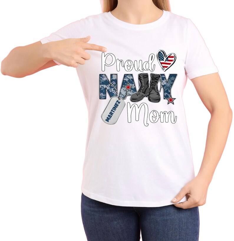 May include: White t-shirt with the text "Proud Navy Mom" in blue and black lettering. The text is surrounded by a camouflage pattern and a red, white, and blue heart. A dog tag with the text "MARTINEZ" is hanging from the bottom of the "Navy" text.