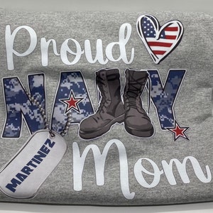 May include: Gray t-shirt with the text "Proud Navy Mom" in white. The word "Navy" is in a blue camouflage pattern with a red star. There are two black boots and a gray dog tag with the text "MARTINEZ" in black.